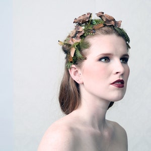 May include: A woman wearing a decorative headpiece made of dried flowers, feathers, and greenery. The headpiece is brown, green, and tan. The woman has fair skin, dark eye makeup, and dark red lipstick. The background is a light, neutral color.