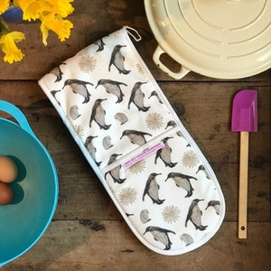 Puede incluir: Un guante de cocina doble blanco con un estampado de pingüinos grises y explosiones doradas. El guante tiene un lazo para colgar y una etiqueta que dice "ART BY CHLOE GARDNER". Una espátula morada y una olla crema también son visibles.