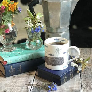 May include: A white ceramic mug with a blue and gray fish design, filled with coffee, sitting on a stack of books. The mug has a handle and is resting on a blue book. There are flowers in vases and a silver coffee pot in the background.