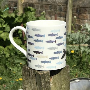May include: White ceramic mug with a pattern of blue and gray fish. The mug has a black fish on the side.