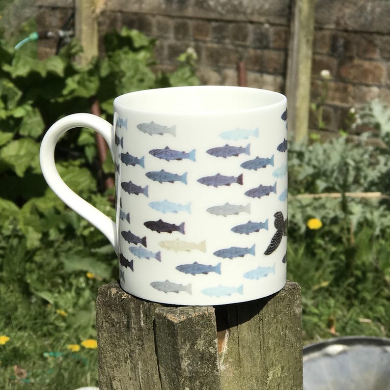 May include: White ceramic mug with a pattern of blue and gray fish. The mug has a black fish on the side.