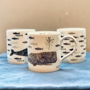 May include: Three white ceramic mugs with a black and white illustration of a whale and fish. The mug in the center has a handle and the whale is in the foreground. The mugs are on a blue fabric surface.