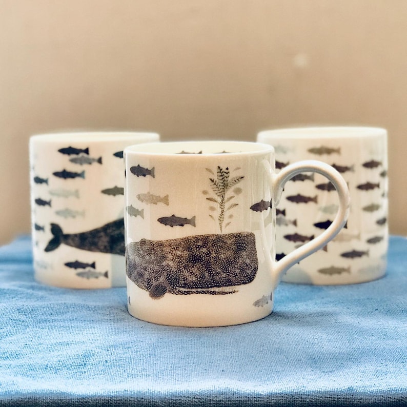 May include: Three white ceramic mugs with a black and white illustration of a whale and fish. The mug in the center has a handle and the whale is in the foreground. The mugs are on a blue fabric surface.