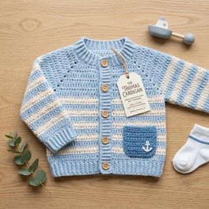 May include: A light blue and cream striped crochet cardigan with long sleeves, a ribbed collar, and four wooden buttons. A pocket features a white anchor emblem. A tag reads The Thomas Cardigan, size 6-12 mos, soft cotton & wool. A white sock is nearby.