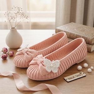 Peut inclure: Une paire de ballerines en tricot côtelé rose clair avec un nœud proéminent sur le bout arrondi. Chaque chaussure est ornée d'un papillon en crochet blanc centré d'une petite perle. Une étiquette en bois marquée MAGIC SHOES est présente.