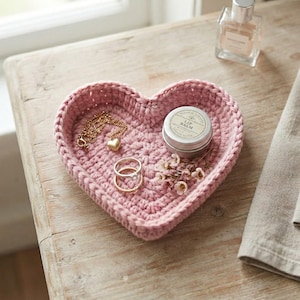 May include: A heart-shaped pink crochet bowl containing a gold-tone chain necklace with a heart pendant, two stacked silver-tone rings, a small round tin of lip balm, and a sprig of small white flowers with light pink centers.