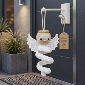 May include: A crocheted angel ornament with a beige head, a gold halo, and white wings attached to a white spiral body. A white cord loop hangs from a white wooden wall hook, which also holds a brown rectangular tag labeled HANDMADE WITH LOVE.