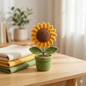Könnte beinhalten: Eine gehäkelte Sonnenblume in einem grünen Topf mit brauner Mitte und gelben Blütenblättern. Die Pflanze hat zwei grüne Blätter an einem zentralen Stiel. Die Sonnenblume ist aus Garn gefertigt und zeigt eine strukturierte Optik mit gleichmäßigen Maschen.