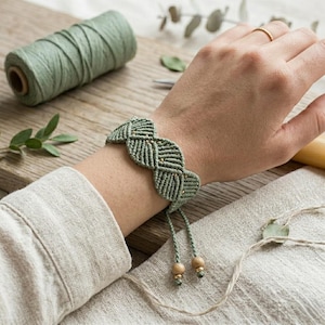 May include: A sage green macrame bracelet featuring a woven wave pattern with small gold-toned beads integrated into the design. The bracelet has two adjustable cord ends, each finished with a single wooden bead at the tip for a secure fit.