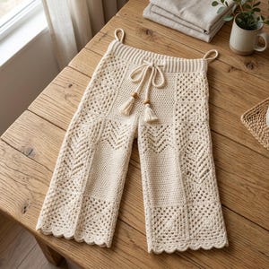 May include: Off-white crochet pants featuring a patchwork design of various stitch patterns, including granny squares and chevron motifs. The pants have an elasticized waistband with a drawstring closure finished with wooden beads and tassels.