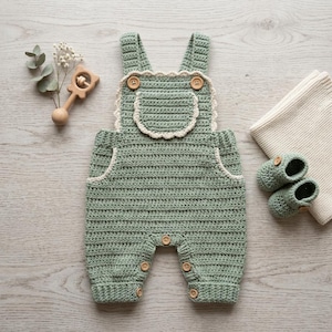 Può includere: Una tutina all'uncinetto color verde salvia con una tasca a pettorina smerlata, due spalline con bottoni in legno e ulteriori bottoni in legno sulle aperture delle gambe. Il set include un paio di scarpine abbinate e un sonaglio in legno.