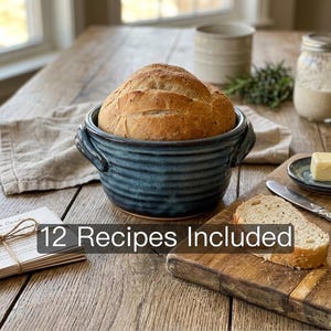 May include: A round, golden-brown loaf of crusty bread sits inside a deep, dark blue ceramic bowl with horizontal ridges and two side handles. The text "12 Recipes Included" is overlaid on the image. A slice of bread rests on a wooden board.