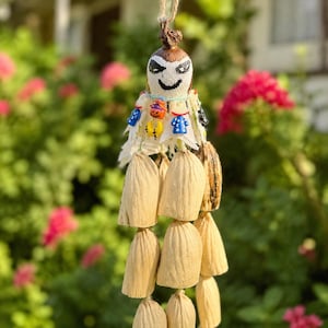 May include: A hanging ornament crafted from dried corn husks, featuring a painted white face with black eyes and a smile. The top is adorned with small, colorful plastic beads shaped like pumpkins and shirts, suspended by a thin twine string.