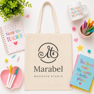 May include: A beige canvas tote bag featuring a black circular logo with the letter M and the text Marabel MOCKUPS STUDIO. The bag is surrounded by a spiral notebook, a white mug, a pink heart-shaped dish, and a blue book with colourful text.