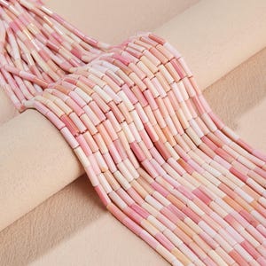 Könnte beinhalten: Mehrere Stränge zylindrischer Perlen in verschiedenen Rosatönen, die von blassem Blush bis zu kräftigem Rosa reichen. Die Perlen haben eine glatte, glänzende Oberfläche und sind in parallelen Reihen angeordnet, was eine gleichmäßige Textur ergibt.