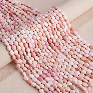 May include: A collection of small, round, flat beads in various shades of soft pink, peach, and cream. The beads are arranged in multiple parallel strands, showcasing a smooth, polished finish and a consistent, uniform shape throughout the set.