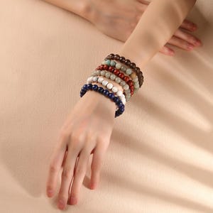 May include: A stack of seven beaded bracelets worn on the wrist. The bracelets feature round beads in various colors including dark blue, white, grey, red, light green, and dark brown, all strung on an elastic cord for a flexible fit.