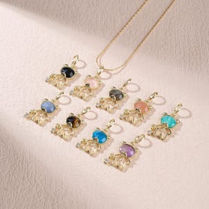 May include: A collection of gold-toned pendants shaped like stylized figures with faceted, multi-colored gemstone heads, including black, pink, grey, blue, tiger eye, orange, turquoise, and purple stones, with one pendant attached to a gold chain.