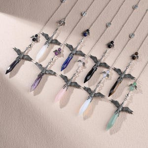 May include: A collection of seven silver-toned metal necklaces, each featuring a pendant shaped like a bird with outstretched wings clutching a faceted, pointed gemstone. The stones vary in color, including dark blue, purple, pink, black, brown, and green.