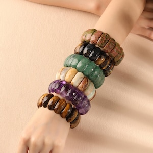 May include: A stack of six faceted gemstone bracelets worn on the wrist. The bracelets feature various natural stones including tiger eye, amethyst, white quartz, green aventurine, black obsidian, and mottled pink and green unakite stone.
