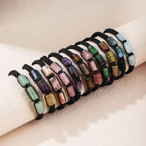 May include: A collection of ten adjustable bracelets, each featuring three rectangular polished gemstone beads in various colors including green, purple, pink, brown, and blue, strung on black cord with a knotted design.
