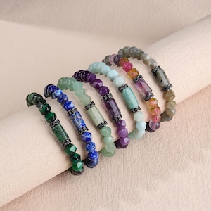 Op de afbeelding: Een collectie van zeven kralenarmbanden met diverse edelstenen in tinten groen, blauw, paars en grijs. Elke armband bevat een centrale cilindrische steenkraal, geflankeerd door twee donkere metalen tussenstukjes en gefacetteerde kralen.