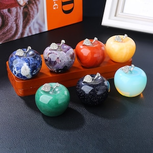 May include: A collection of seven polished stone apple figurines in various colors including blue, purple, orange, yellow, green, dark blue with glitter, and translucent light blue, each topped with a silver-toned metal leaf with crystals.