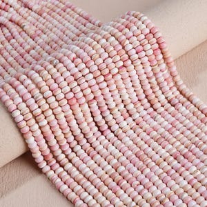 May include: Multiple strands of small, round, disc-shaped beads in varying shades of light pink, soft rose, and creamy white. The beads have a smooth, matte finish and are arranged in parallel rows, showcasing a consistent size and shape.