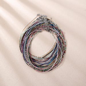 Op de afbeelding: Een verzameling van meerdere gelaagde kettingen gemaakt van kleine, veelkleurige gefacetteerde edelsteenkralen. Elke ketting bestaat uit een dunne streng van kleine, gepolijste steentjes in diverse tinten zoals blauw, roze, groen en wit, afgewerkt met een zilveren sluiting.