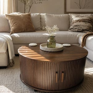 May include: A round, dark wood coffee table featuring a slatted tambour door design with two vertical rectangular handles. On top sits a circular, ribbed light-colored tray holding a small, clear green glass vase filled with white flowers.