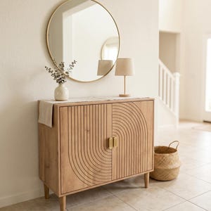 May include: A light wood cabinet with two doors featuring carved vertical and arched lines, gold rectangular handles, and four tapered legs. A round gold-framed mirror, a small white vase with eucalyptus, and a beige lamp sit on top.