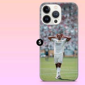 May include: A clear smartphone case featuring a printed image of a soccer player in a white jersey with the text Emirates Fly Better, standing on a green field with hands cupped behind ears. The case has a cutout for a triple-lens camera system.