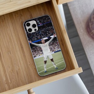 May include: A rectangular smartphone case featuring a printed image of a soccer player in a white uniform standing on a green field with arms outstretched. The player wears a white jersey with the text Emirates, white shorts, and white socks.