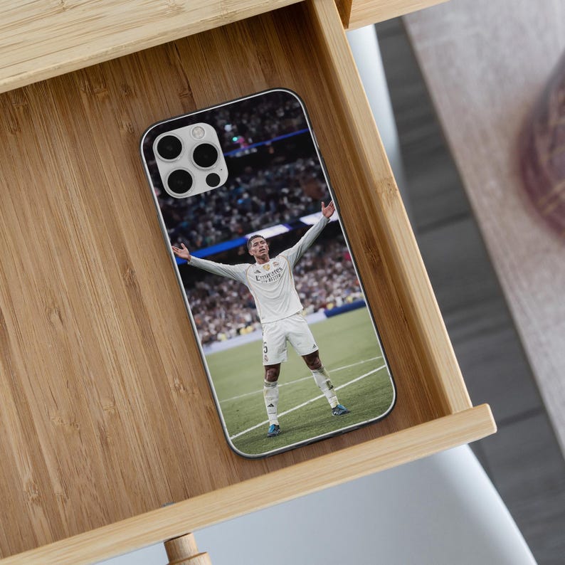 May include: A rectangular smartphone case featuring a printed image of a soccer player in a white uniform standing on a green field with arms outstretched. The player wears a white jersey with the text Emirates, white shorts, and white socks.