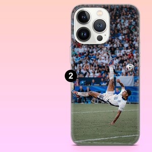 May include: A clear smartphone case featuring a printed graphic of a soccer player in a white uniform performing a bicycle kick on a green field. The player is mid-air with one leg extended upward toward a soccer ball, with a blurred stadium crowd.