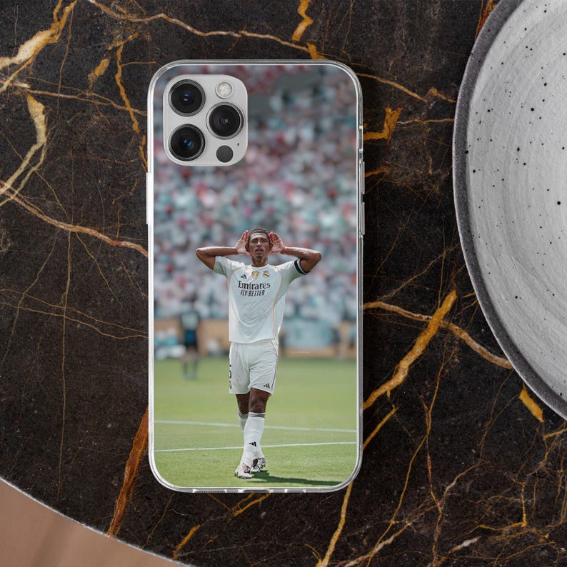 May include: A clear phone case featuring a printed image of a soccer player in a white Real Madrid jersey with the text Emirates Fly Better, standing on a green field with hands cupped behind ears in front of a blurred stadium crowd.