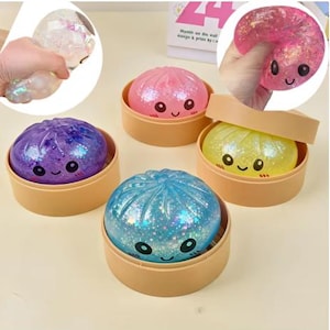 Puede incluir: Una colección de bolas antiestrés coloridas y llenas de purpurina con forma de empanadillas, cada una con una cara sonriente. Las empanadillas están en recipientes de color beige. Los colores incluyen rosa, azul, morado y amarillo. Una está siendo apretada por una mano.