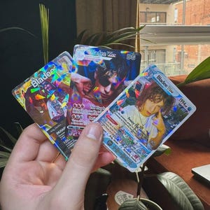 May include: Three rectangular trading cards featuring holographic foil details and portraits of the artist Bladee. The cards display text including Bladee, VMAX, DG, 1994, Frosty The Snowman, and various game statistics printed in colorful fonts.