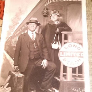 May include: A black and white vintage photograph of a couple standing on a train platform. The man is wearing a suit and tie, and the woman is wearing a coat and hat. The train is in the background, and there is a sign that says "Long Beach" on the side of the train.