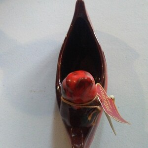 May include: A ceramic canoe-shaped planter in shades of brown and red. A red figure is inside the canoe, and a tag is attached. The planter is likely for small plants or decorative purposes.