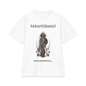 May include: A white short-sleeve t-shirt featuring a central graphic of an alien figure with a split design, surrounded by thorny branches and a celestial sphere. The text PARANORMAL? appears above the graphic and EXTRATERRESTRIAL. below it.