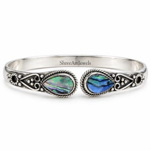 Op de afbeelding: Een zilveren armband met twee druppelvormige inlays van abalone-schelp in iriserende groene en blauwe tinten. De metalen band is voorzien van gedetailleerde touwranden, kraalaccenten en krulwerk. De tekst ShreeArtJewels is duidelijk zichtbaar.