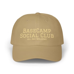 Może przedstawiać: Beżowa czapka z daszkiem z napisem BASECAMP SOCIAL CLUB wykonanym jasnokremową czcionką blokową, z mniejszym napisem ALL ARE WELCOME umieszczonym bezpośrednio pod spodem w tym samym jasnokremowym kolorze, wyśrodkowanym na przodzie czapki.