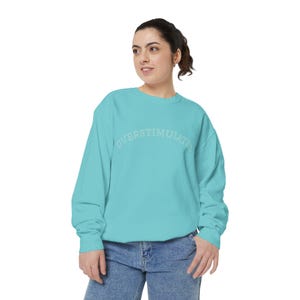Peut inclure: Un sweat-shirt à col rond et manches longues de couleur turquoise vif, arborant le mot OVERSTIMULATED imprimé en lettres claires et arquées sur la poitrine. Le vêtement présente un col, des poignets et un ourlet côtelés, avec un jean bleu.