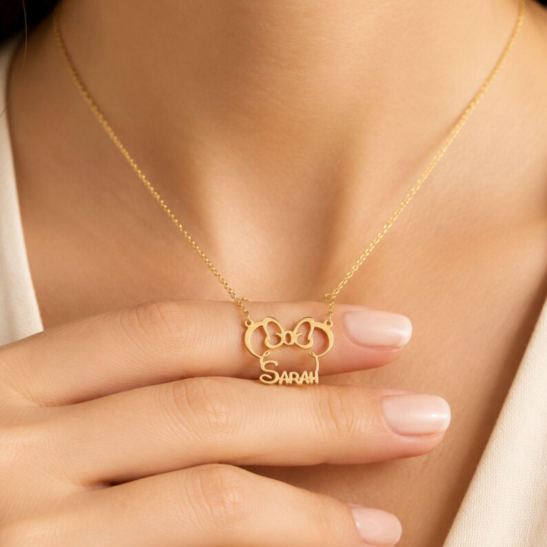 May include: A gold-toned necklace featuring a pendant shaped like a stylized mouse head with a bow at the top. The name SARAH is integrated into the lower portion of the pendant design. The pendant is attached to a delicate, thin gold-toned chain.