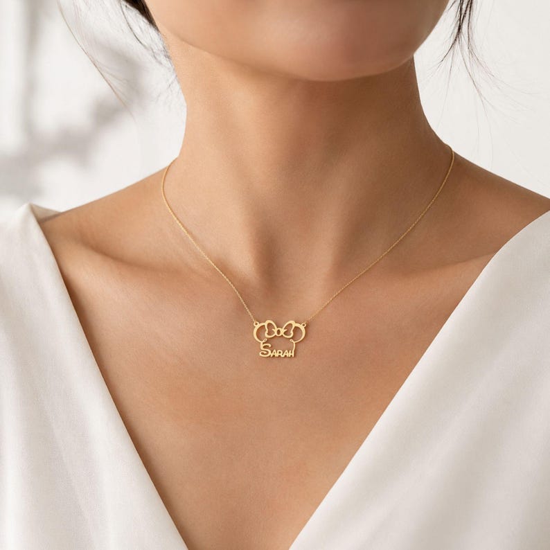 May include: A gold-toned necklace featuring a delicate chain and a central pendant shaped like a stylized mouse head silhouette with a bow, incorporating the name Sarah in a cursive script font integrated into the lower portion of the design.