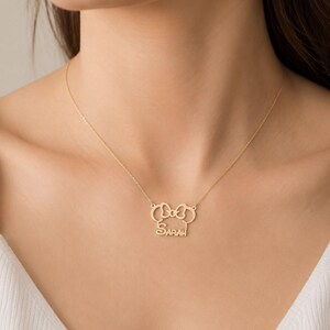 May include: A gold-toned necklace featuring a pendant shaped like a stylized mouse head with a bow at the top and the name SARAH written in capital letters across the center. The pendant is attached to a delicate gold-toned chain.