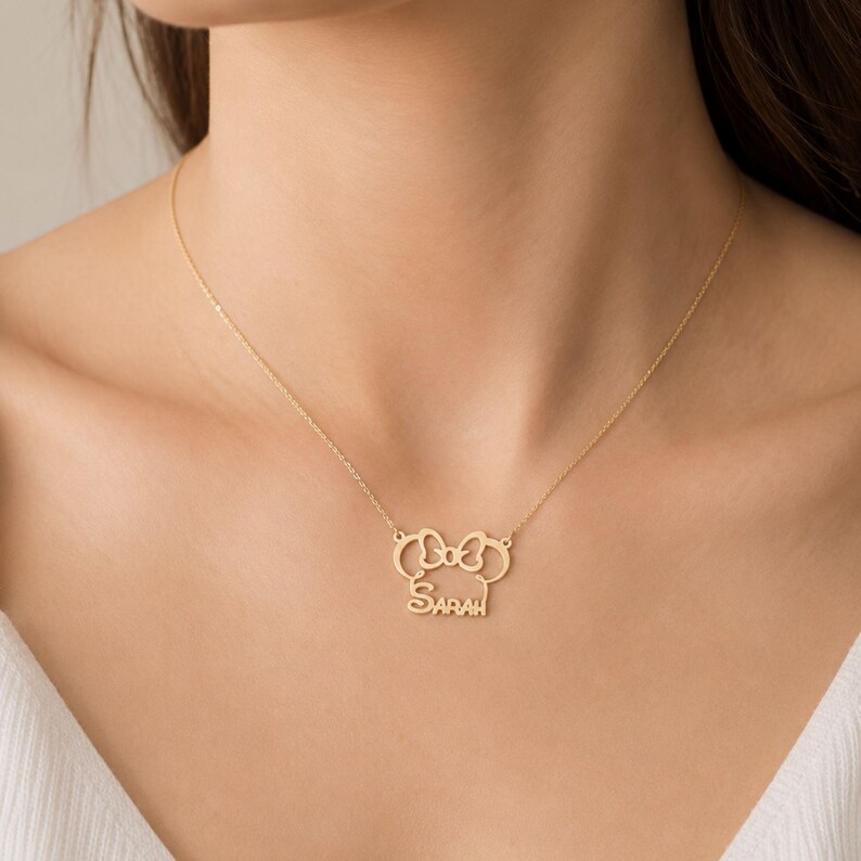 May include: A gold-toned necklace featuring a pendant shaped like a stylized mouse head with a bow at the top and the name SARAH written in capital letters across the center. The pendant is attached to a delicate gold-toned chain.