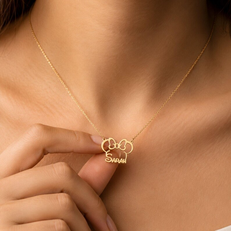May include: A gold-toned necklace featuring a pendant shaped like a stylized mouse head silhouette with a bow at the top and the name Sarah written in cursive script integrated into the lower portion of the design, suspended on a thin chain.