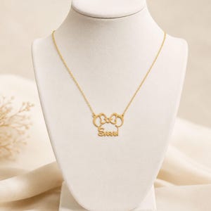 May include: A gold-toned necklace featuring a pendant shaped like a stylized mouse head with a bow at the top and the name Sarah written in cursive script across the bottom, suspended from a delicate matching gold-toned chain on a display bust.